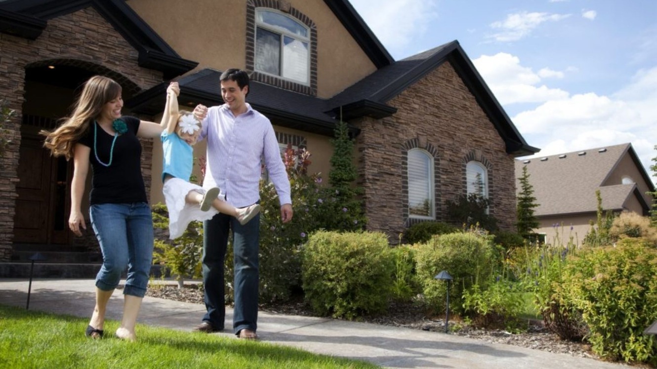 A family of three (mother, father, and daughter) are walking in the grass. Behind them is their house. The mother and father are playfully swinging the daughter. They are happy.